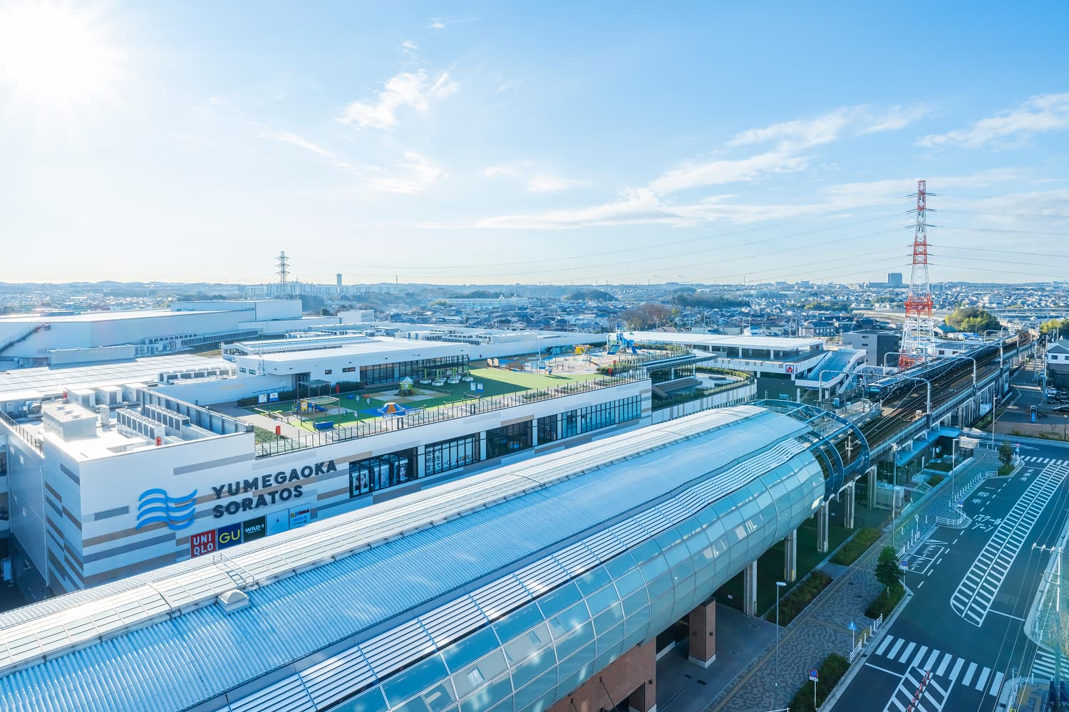 手前に YUMEGAOKA SORATOS と書かれた大型商業施設、隣にガラスドームの駅舎と線路、奥に街並みが広がる鳥瞰写真、イメージ